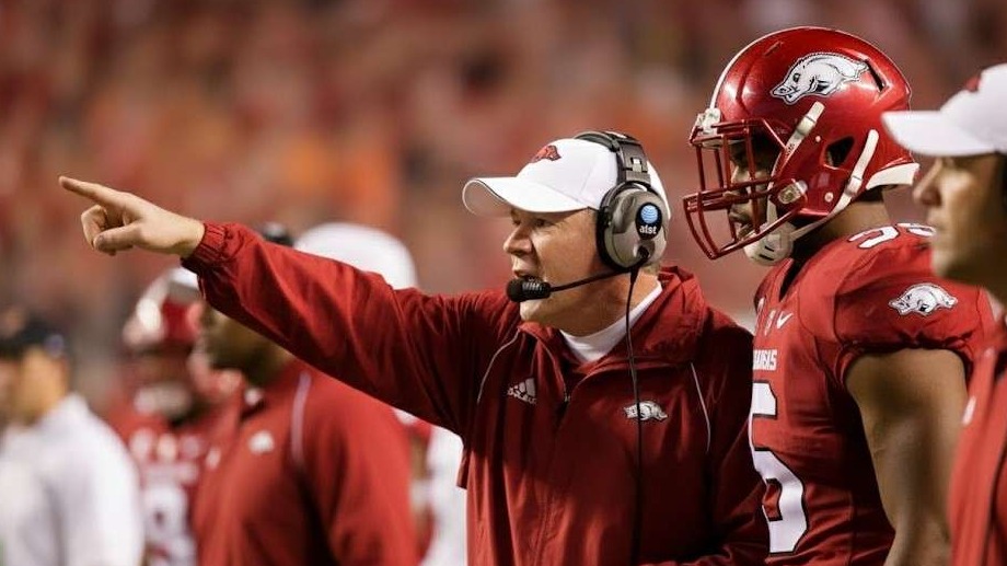 Bobby Petrino speaking at a sports press conference with team logos.
