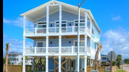 Beautiful coastal vacation home exterior, sunbathed house with wraparound porch, welcoming for Oak Island vacation, coastal color palette, fluttering flags, and swaying palms in the background