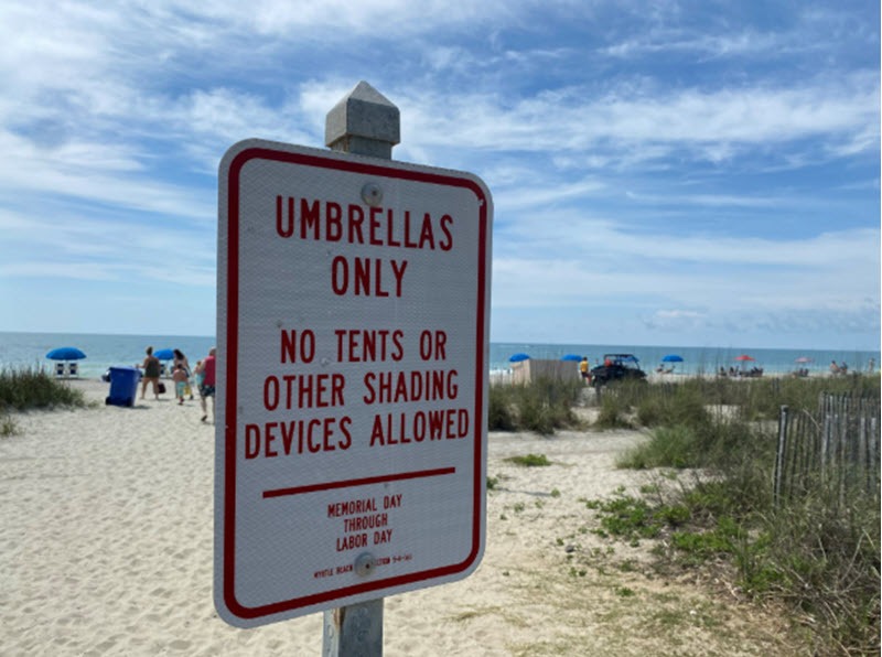 smiling beachgoers reading posted umbrella and tent rules at a Myrtle Beach entry sign children with sand toys lifeguard tower background vibrant community feel late afternoon sunlight