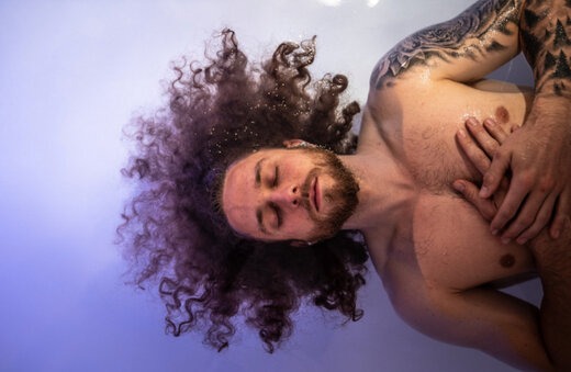 A man with long curly hair and a tattooed arm floats peacefully in a sensory deprivation tank, eyes closed and hands resting on his chest, surrounded by calming purple-blue light.