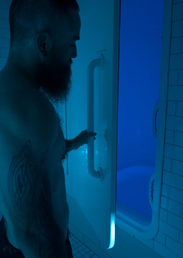 A bearded man with a tattoo on his arm stands in dim blue lighting, opening the door to a sensory deprivation float tank in a tiled wellness facility.