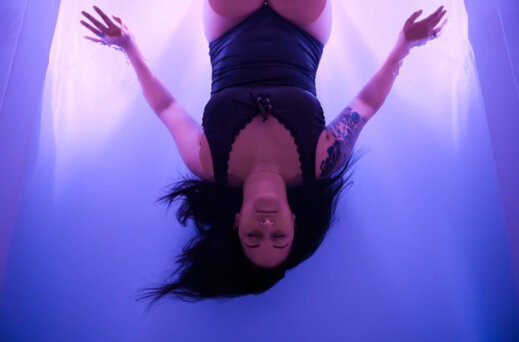 A woman in a black swimsuit floats effortlessly in a sensory deprivation tank, eyes closed and arms relaxed, surrounded by soft purple lighting that enhances the calm atmosphere.