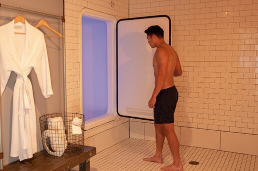 A man in black swim shorts stands in a private spa room, preparing to enter a sensory deprivation tank with soft purple lighting; a robe and towels are neatly arranged nearby.