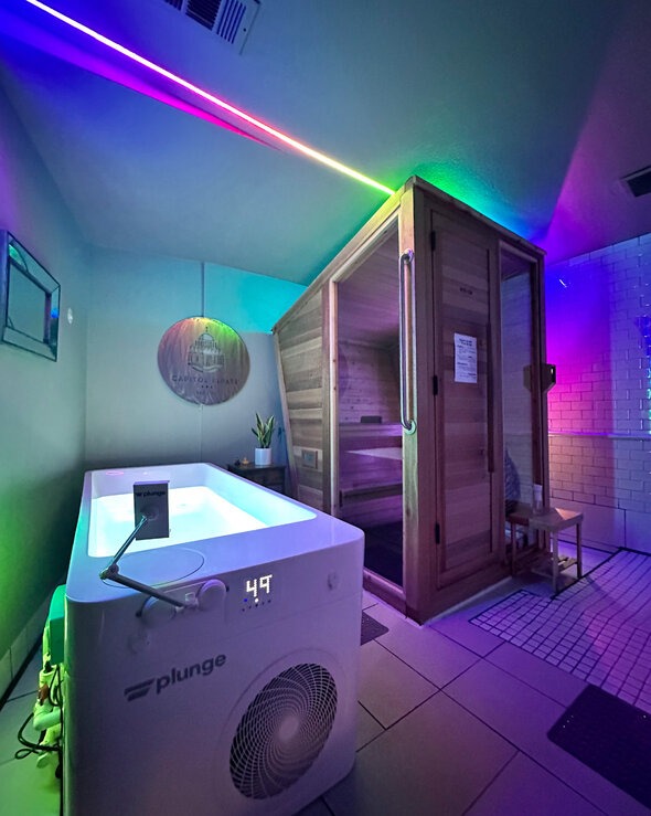 A modern wellness room features a cold plunge tub displaying a temperature of 49°F and a cedar infrared sauna, illuminated by vibrant LED mood lighting across the ceiling.