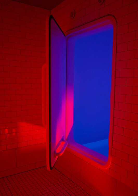 A sensory deprivation float tank glows with deep blue light inside a red-lit tiled room, with the tank door open and inviting, creating a striking contrast of colors.