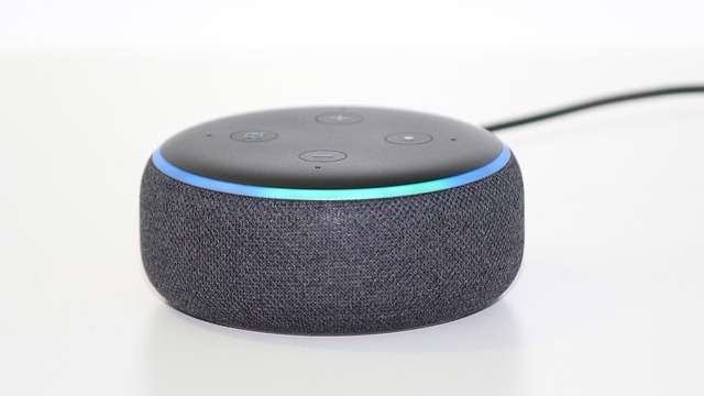 White smart plug on a wooden table, blurred smart speakers behind.