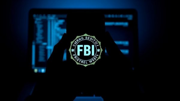 Silhouetted hand holds phone with FBI seal over laptop - fake federal agent scams.