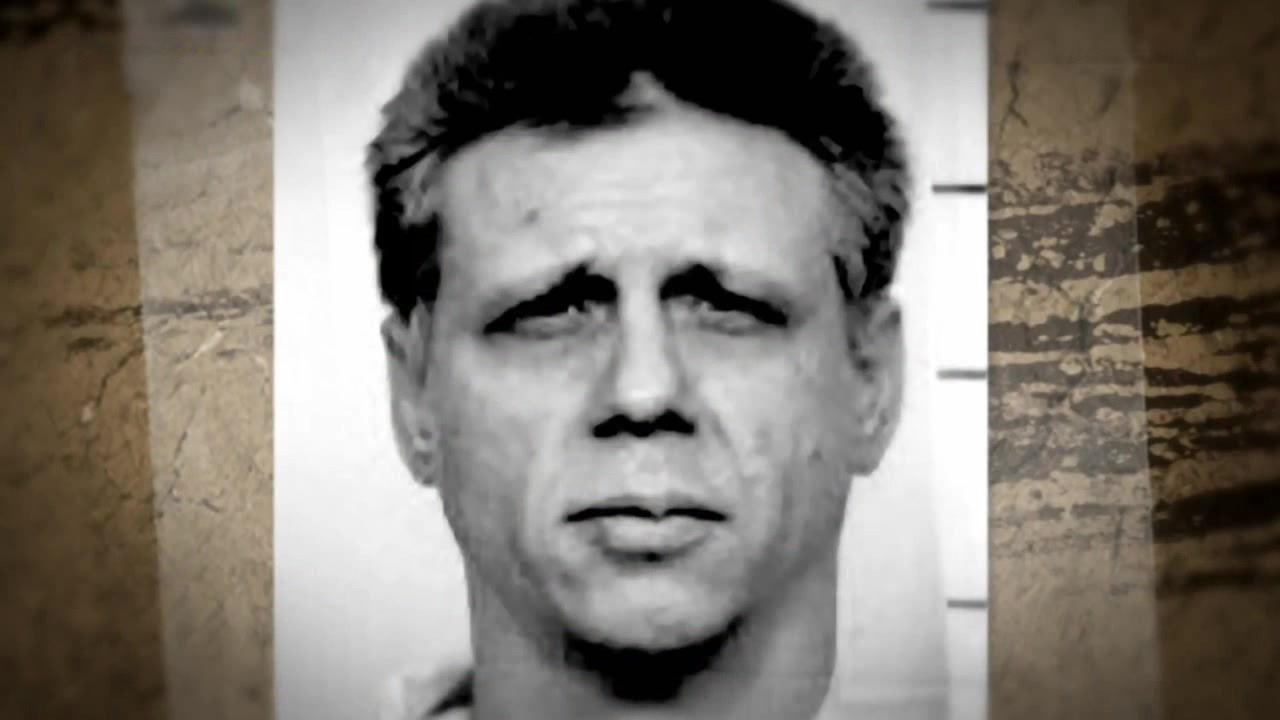 Older man in prison visiting room, solemn expression, white uniform.