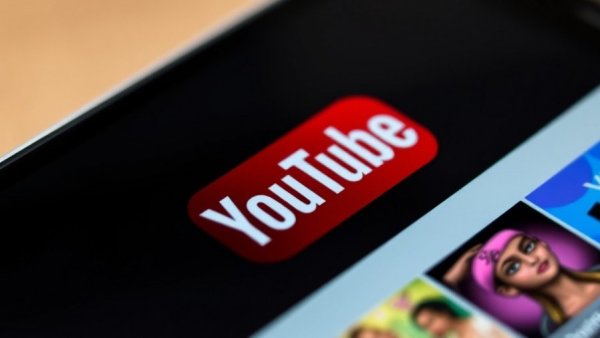 YouTube logo on smartphone with blurred background displaying site thumbnails, Do Faces Help YouTube Thumbnails.
