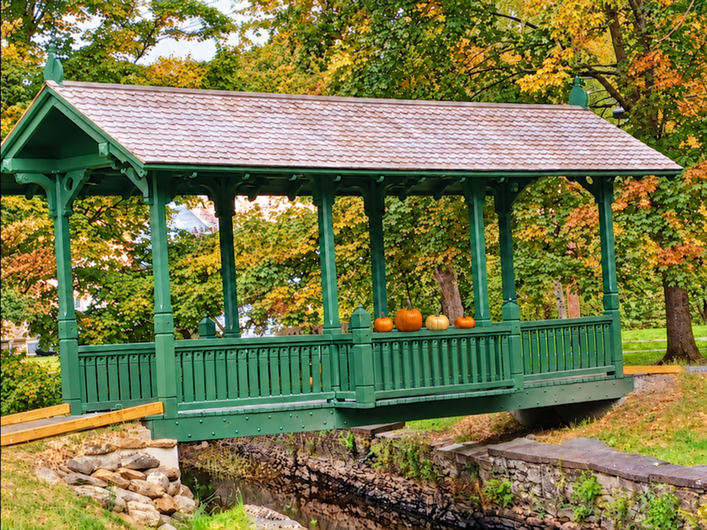 Green covered footbridge in Cambridge, decorated with a small row of pumpkins on its railing and surrounded by early autumn foliage.