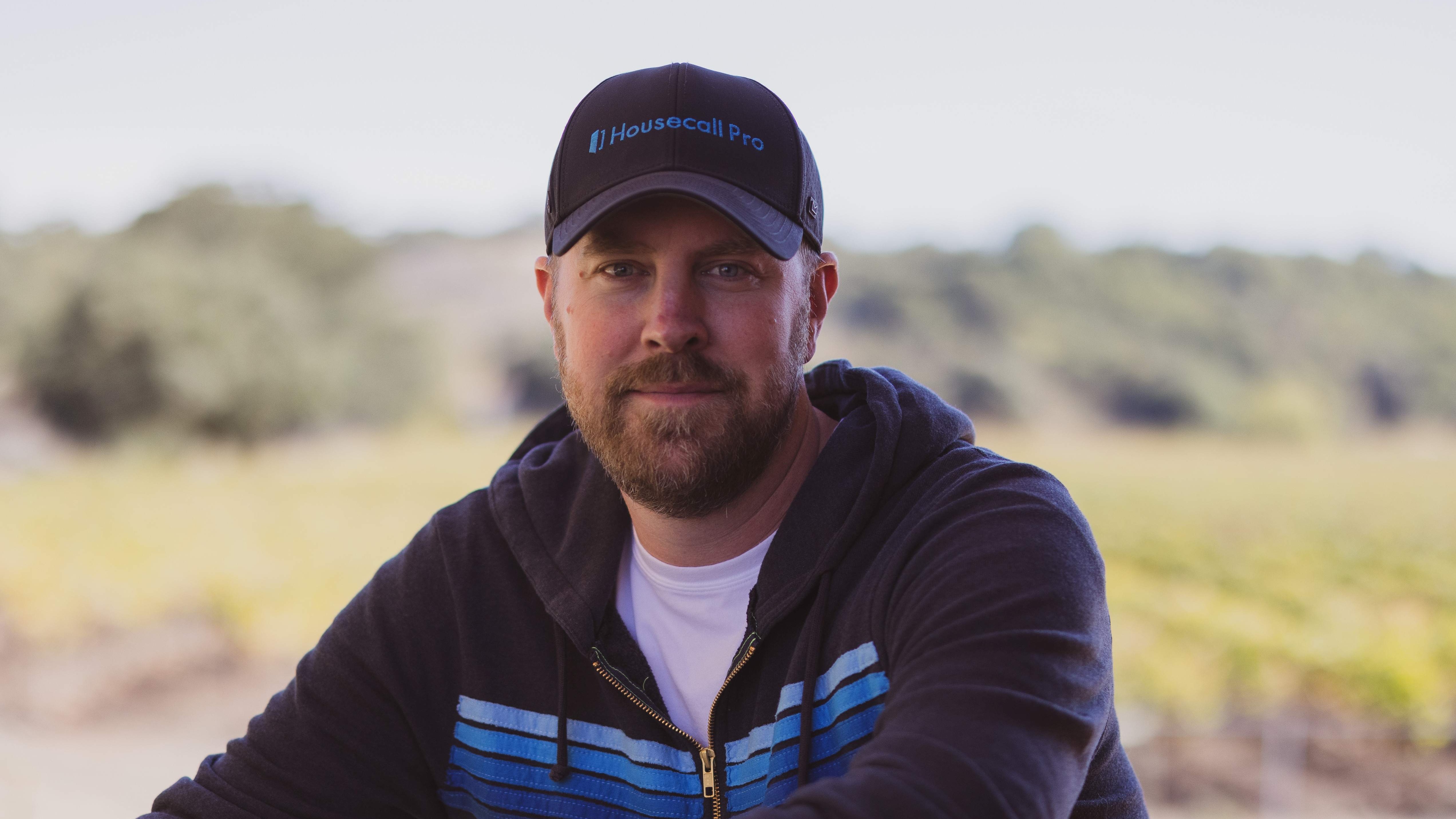 Man wearing hoodie and cap sitting outside, mild smile, blurred landscape.