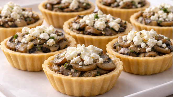 Mushroom and goat cheese tartlets in golden mini tart shells, topped with crumbled goat cheese and served on a white platter.
