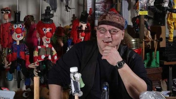 Older man in workshop holding Müll-Marionetten puppet with colorful details.