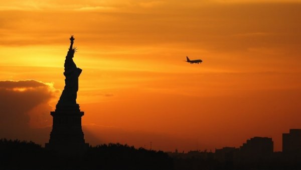 Dramatic view of Statue of Liberty silhouette with distant airplane at sunset.