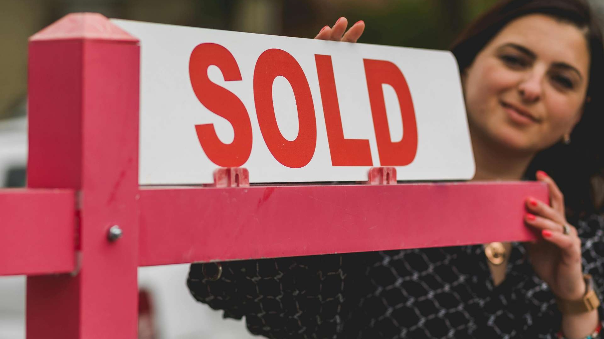 A person partially visible behind a pink fence holds up a white sign with large red letters reading “SOLD.”