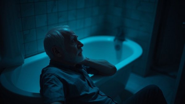 Contemplative older man in a dimly lit bathroom with soft blue lighting.