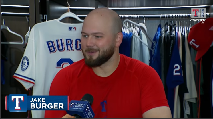 Baseball player smiles in locker room, jerseys hang behind.